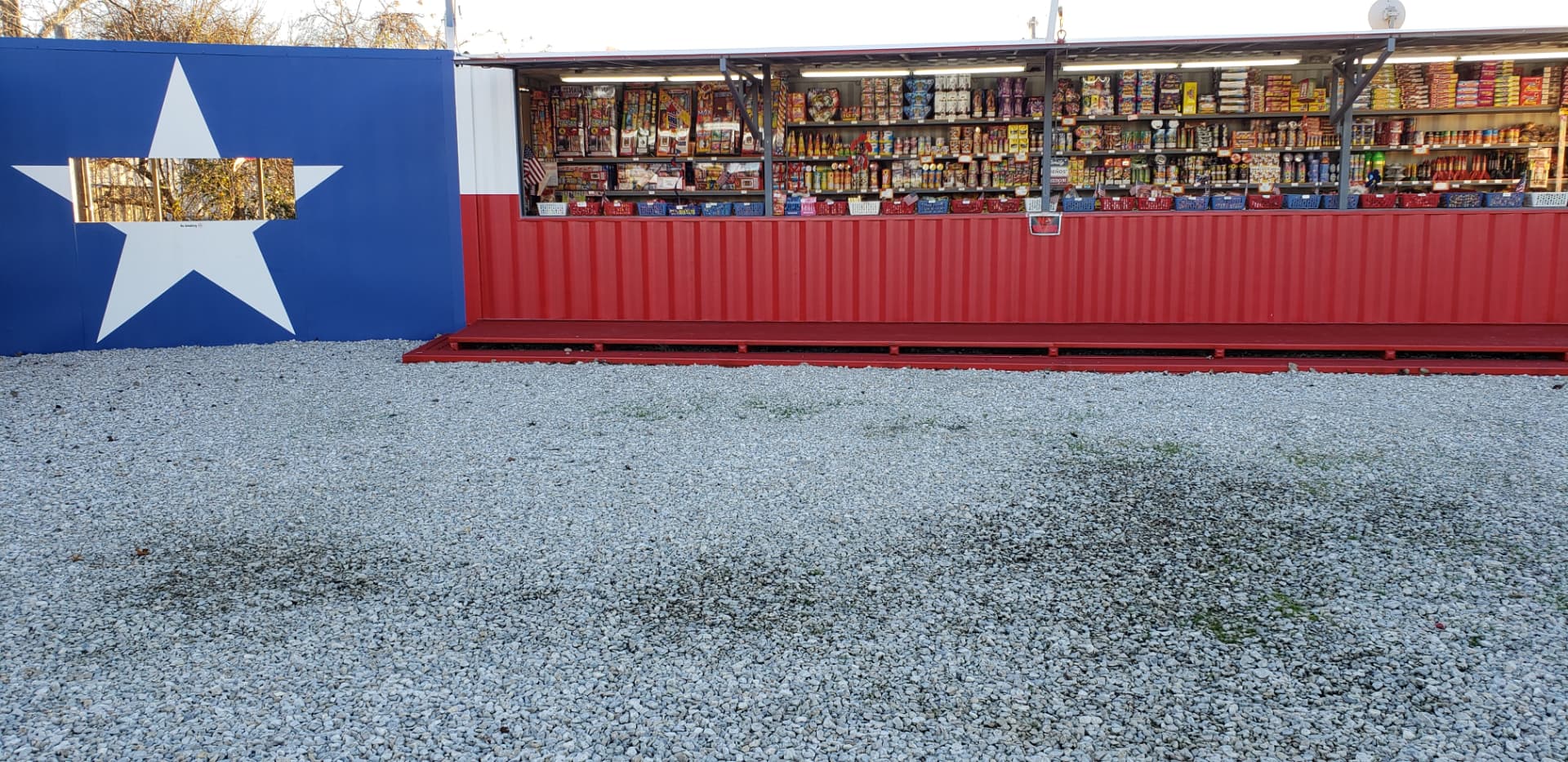 The All About Pyrotechnics stand in daylight, shelves packed with fireworks beside the lone star wall
